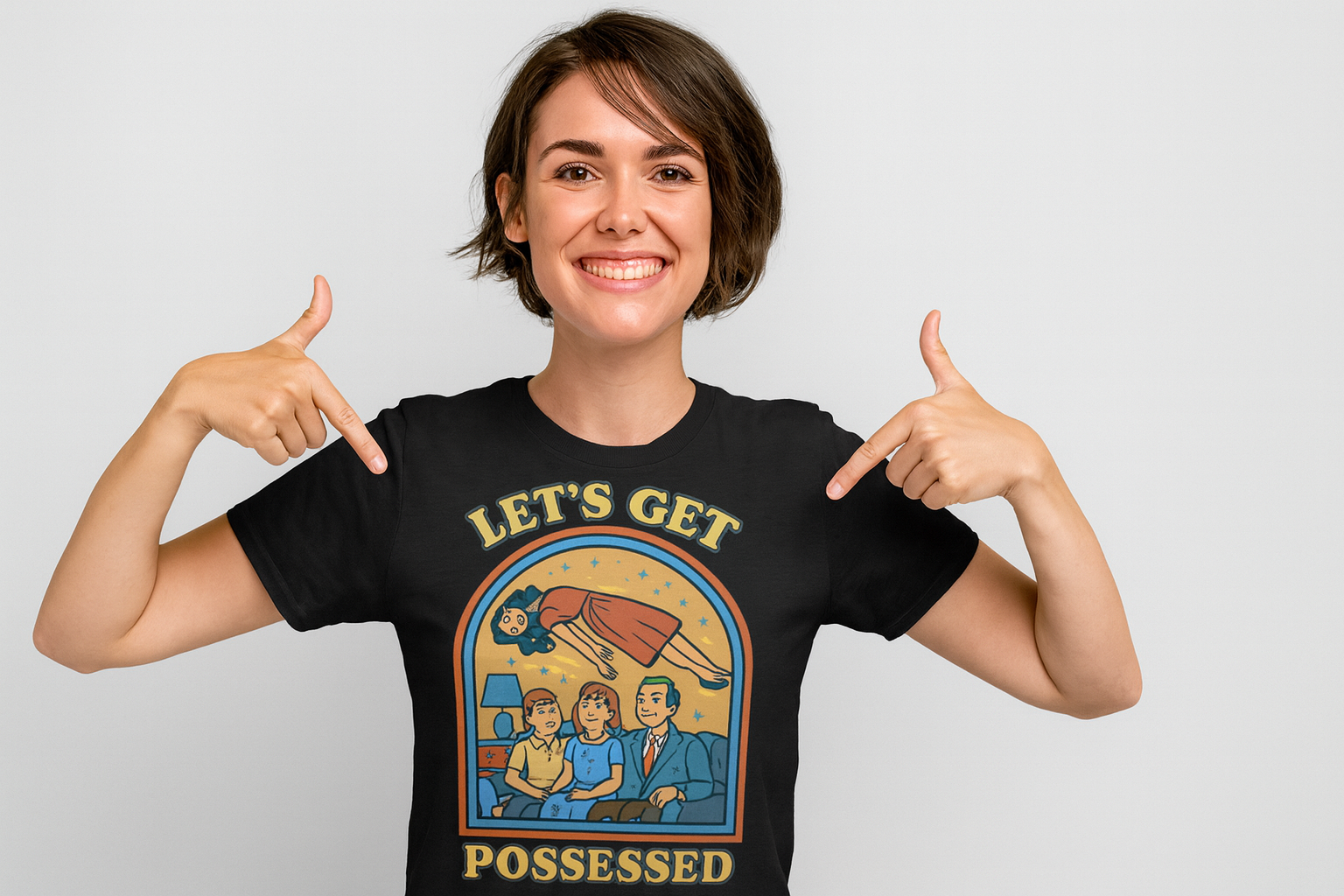 Joven de unos 30 años, sonriente y feliz, con camiseta negra con diseño retro 'Let's Get Possessed'. Señala hacia la cámara con ambas manos, fondo gris claro