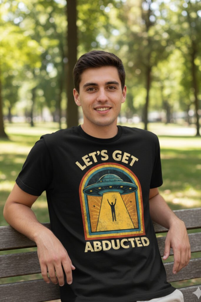 Camisetas frikis con humor Hombre joven sentado en un banco del parque con camiseta friki con humor ‘Let’s Get Abducted’, diseño retro de ovni y abducción