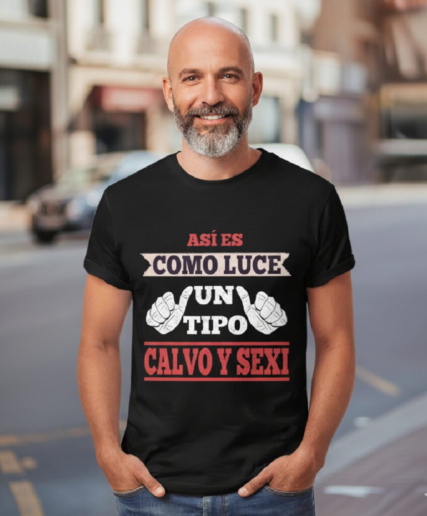 Un hombre calvo de unos 50 años, con una barba gris bien cuidada, mira a la cámara con una sonrisa amable. Viste una camiseta negra con el mensaje "ASÍ ES COMO LUCE UN TIPO CALVO Y SEXY", presentado con un diseño distintivo que incluye dos pulgares hacia arriba. El hombre tiene las manos metidas en los bolsillos de sus pantalones vaqueros, y está de pie en un entorno urbano con edificios y un vehículo ligeramente desenfocados al fondo.